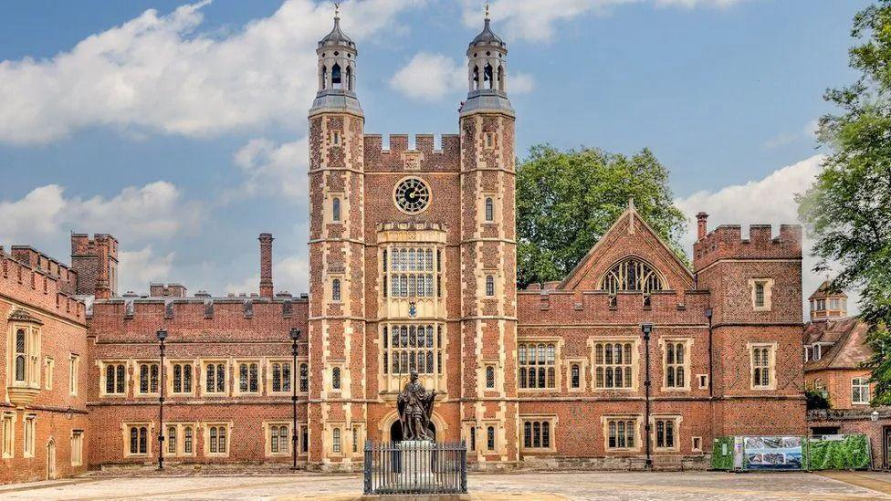 An Eton College building constructed of red brick with a clock tower in the middle and a black statue on a white plinth in front of the entrance.