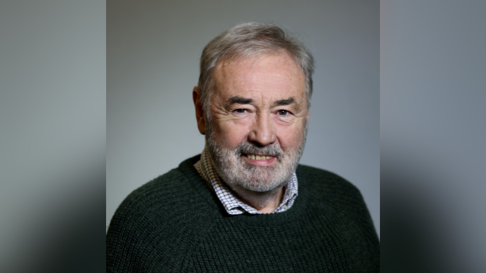 John Allen. He has white hair and beard and brown eyes. He is wearing a green jumper with check shirt underneath.