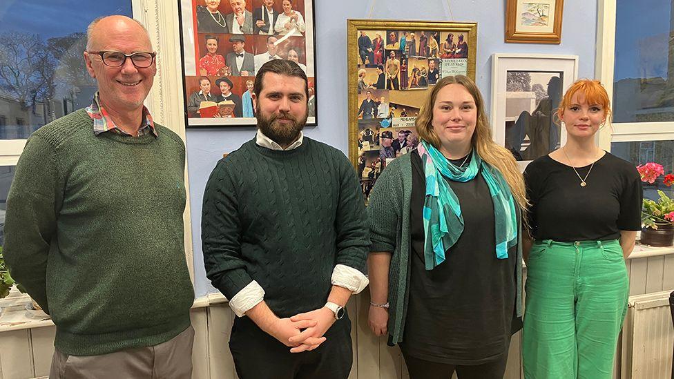 Four members of the Power Hull group stand by a wall inside the Lonsdale Community Centre. From left to right is Kevin Paulson who has short white hair and weras glasses an a green sweater, Ryan Ward who has black hair and beard and wears a dark green jumper, Ellen Clarke who has long brown hair and wears a blue patterned scarf, black shirt and green cardigan and Lauren Duguid who has short red hair a black top and green trousers.