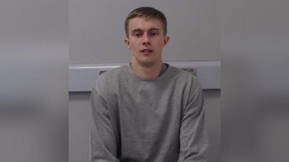 A young man with short brown hair, pictured in a grey sweatshirt against a grey wall