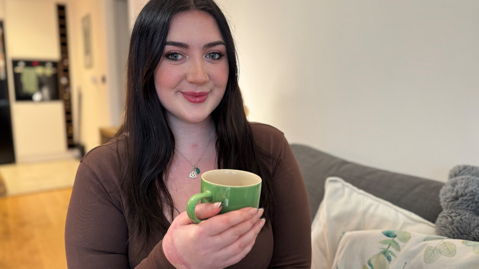 Becca Heritage, a young woman with black hair who is wearing a brown top, holding a green mug and smiling at the camera