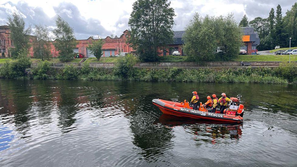 A rescue lifeboat moves along the still waters of a river past some industrial buildings in the background