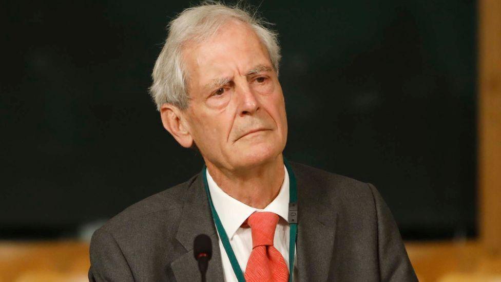 Sir Mark Jones, with grey hair, looks to his left while sitting in front of a dark background. He is wearing a dark grey suit, a white shirt and an orange tie. 