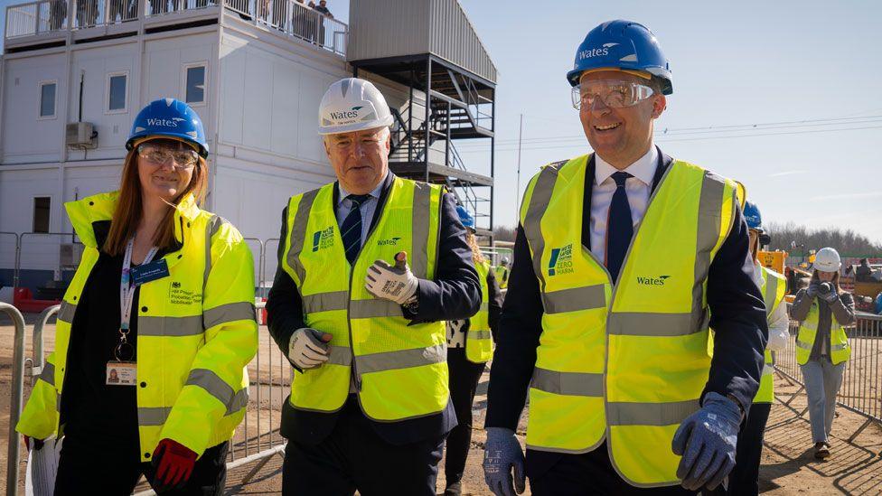 A woman and two men wearing hard hats and high-vis yellow jackets over their dark clothing. They are walking along with a white two-storey container block behind them. The man on the right is Lord Timpson.