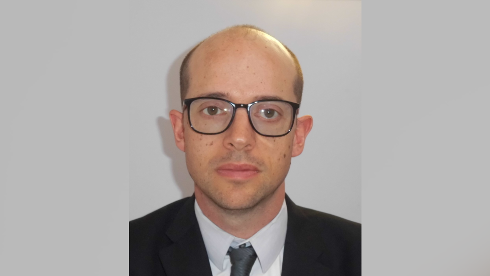A police head shot of a bald man wearing black-rimmed glasses. He is wearing a black suit, a white shirt and a grey tie.