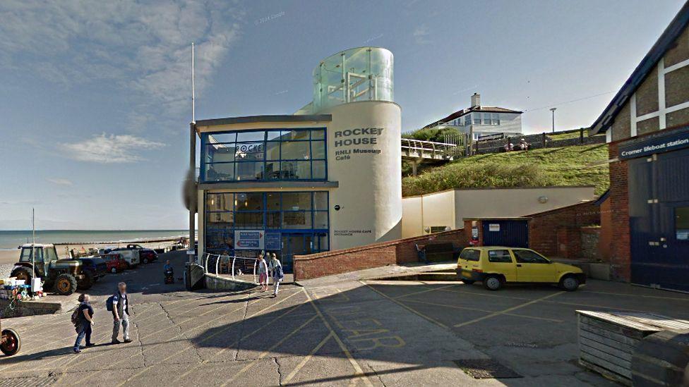 The exterior of the museum and cafe, situated off the gangway to Cromer beach. The museum has a section that is like a lighthouse turret, with a glass viewing point. Beside it is the entrance to the lifeboat station. It is a sunny day and there are a number of people walking outside and there are cars and a tractor parked outside.