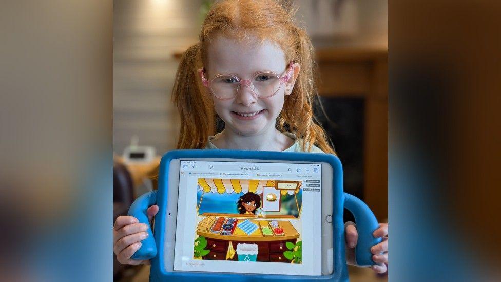 A young girl with ginger hair and glasses holds a tablet computer towards the camera - it has a game on it with a girl sitting at a table.