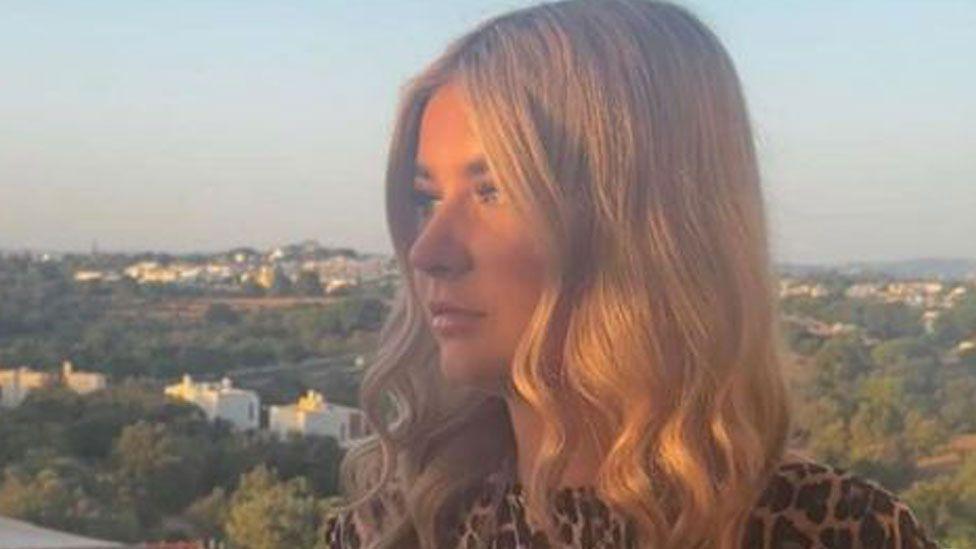 Abi Quinlisk, who has shoulder-length blonde hair and is looking off to the left. She is wearing a brown and cream top. Behind her is a scene showing trees and white houses.