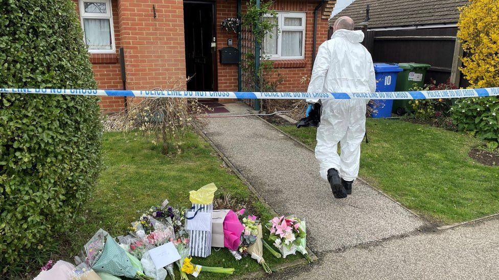The garden of a brick-built house with bushes to the side and small grass lawns either side of a stone path. There is police tape across the garden, and a person in a white hazmat overall is walking towards the front door. There are bouquets of flowers on the lawn to the left.