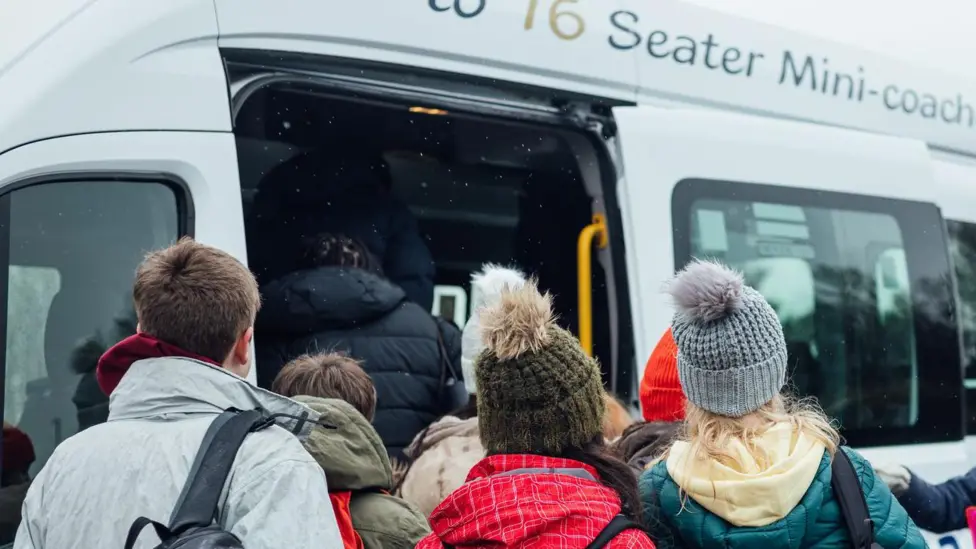 A group of children in coats and woolly hats, some carrying rucksacks, stand in front of a minibus. Their backs are to the camera.