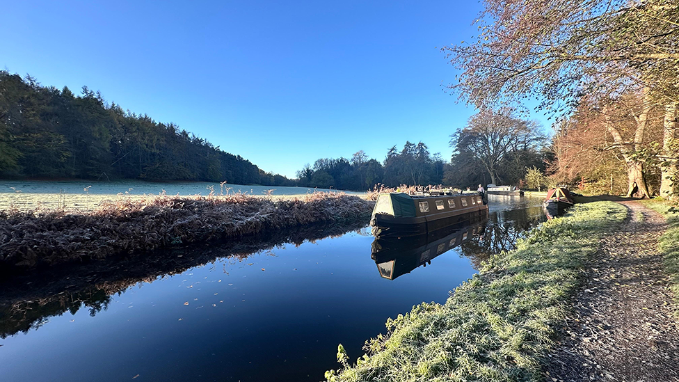 A narrowboat on a canal in Kinver. There is a path and trees to the right and a field with trees in distance on the left