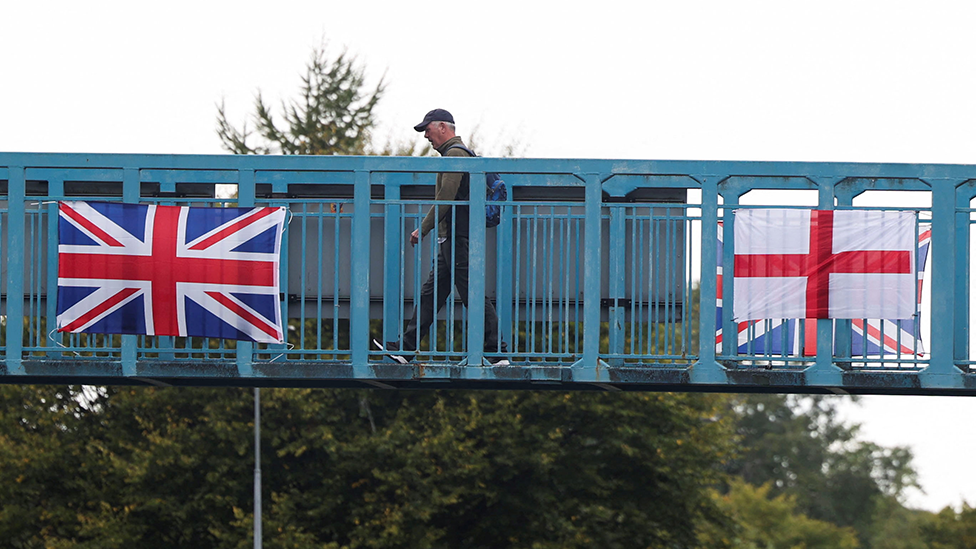 A blue footbridge with a union jack and England flag attached to it. A man is walking over it, while there are trees in the background
