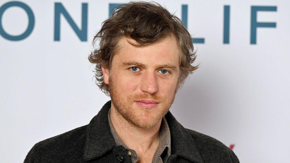 Johnny Flynn standing looking at the camera against a white backdrop at a film premiere