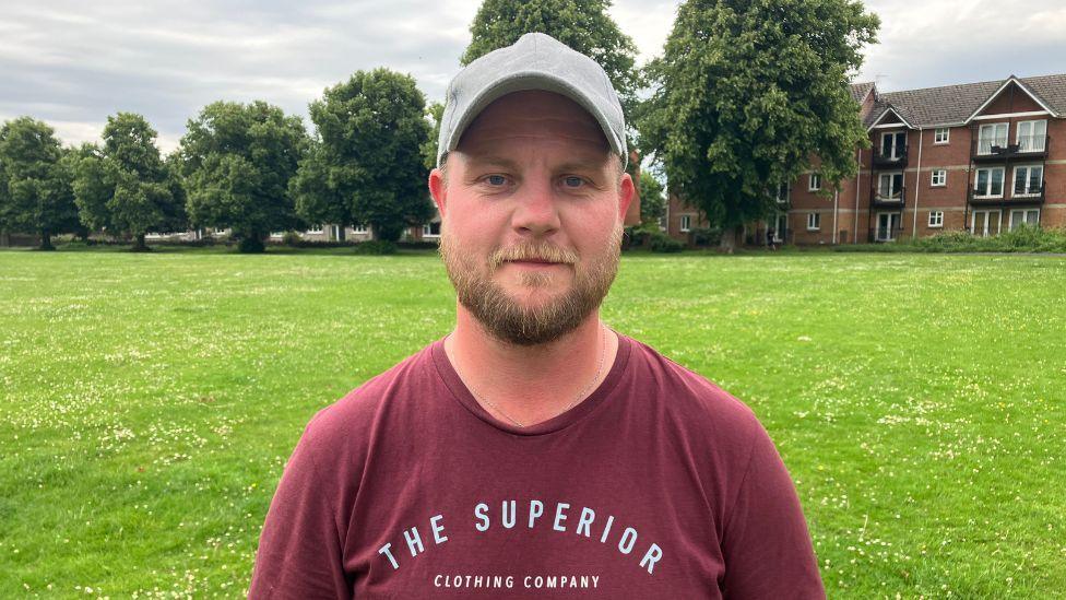 Rhys looks at the camera. He is wearing a burgundy t-shirt and has a grey baseball hat on. He has a short sandy-coloured beard. Grass can be seen behind him with a block of flats in the background.