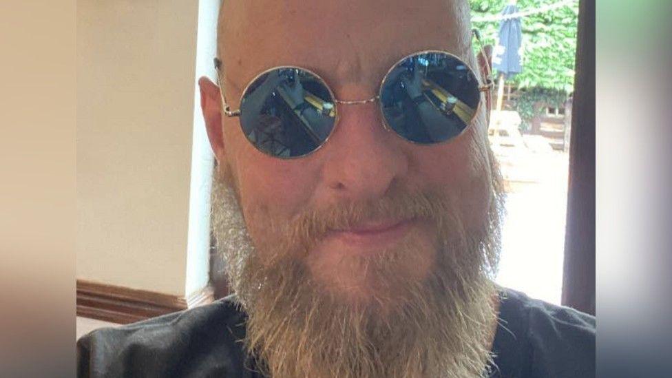 A close-up of Paul Hart, who is bald and has a long beard and is wearing sunglasses. He is in front of a window and a parasol and garden furniture can be seen outside.