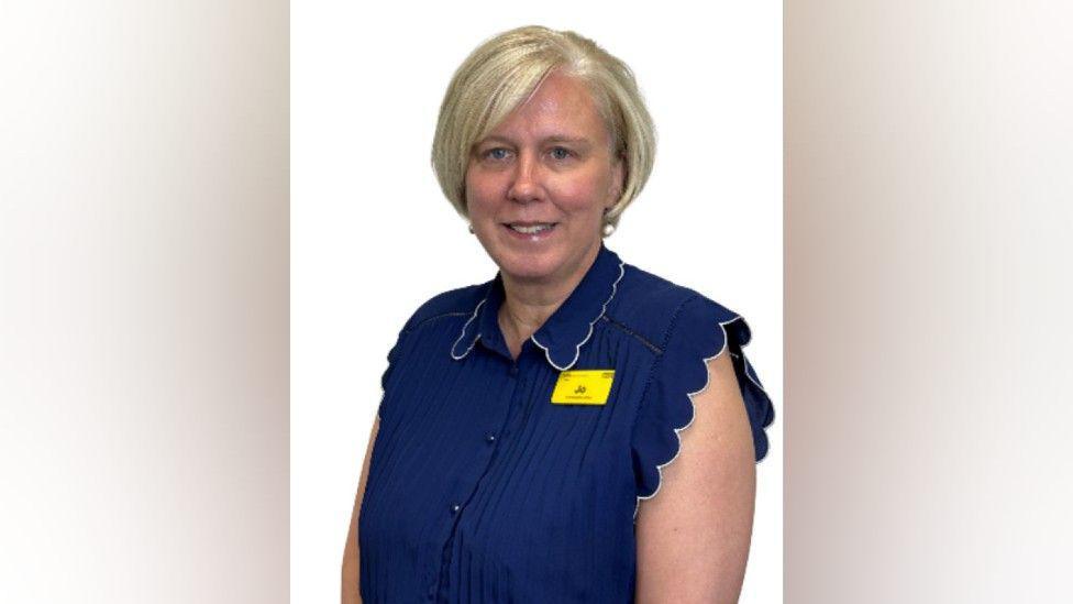 A woman with a short white blonde bob hairstyle is wearing a navy blue blouse and pearl earrings. She has a yellow name badge on that reads "Jo".