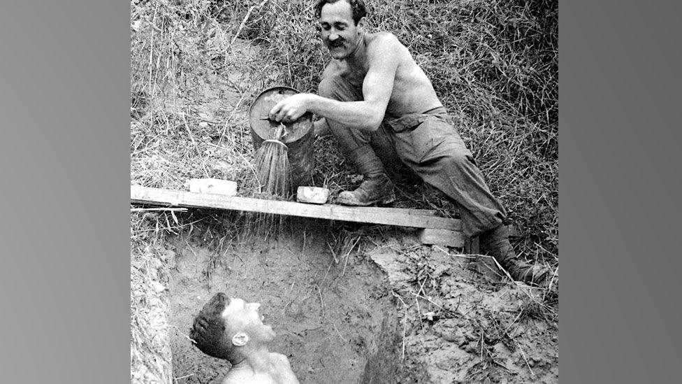 A black and white World War Two photo in Burma. It shows a bare chested Wing Cdr Lander on the right, resting on a plank over a hole and pouring water from a watering can onto a man below who is laughing up at him. 