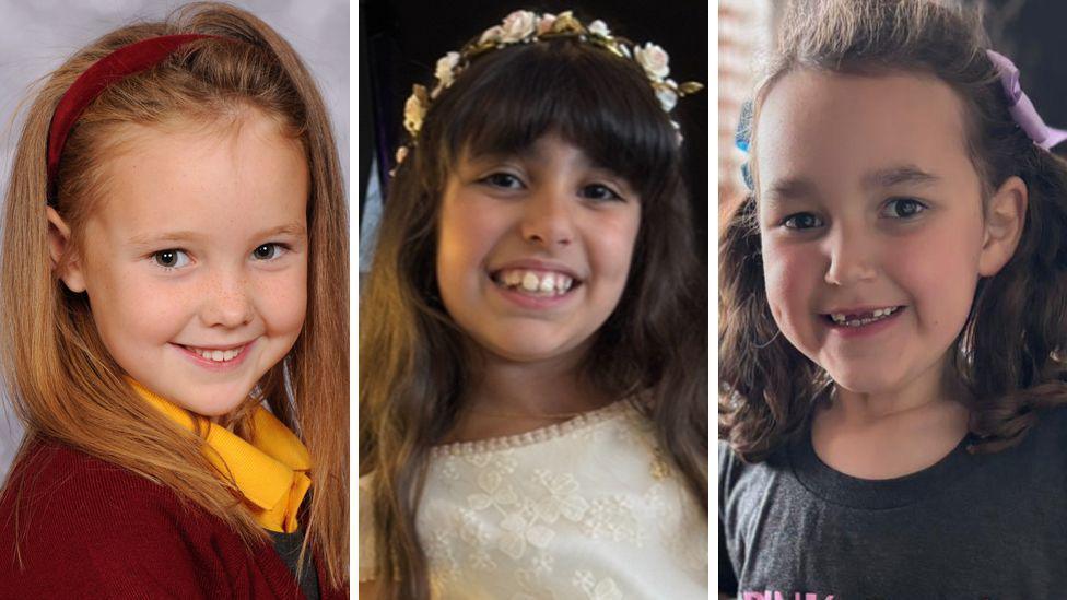 Elsie Dot Stancombe is pictured in school uniform - with maroon cardigan, yellow shirt and grey pinafore dress and matching maroon hairband. Alice da Silva Aguiar is wearing a white lace dress and floral headband, and Bebe King has her hair in bunches and ribbons and is wearing a black T-shirt. All three are smiling at the camera. 