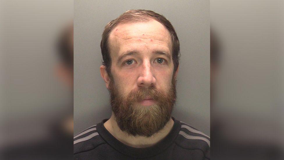 Police custody image of Lee Merrison he has short brown hair and a beard and is wearing a dark shirt with grey stripes on the shoulders