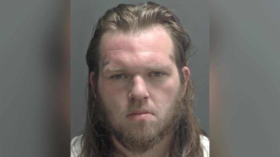 An expressionless man stares at the camera in a police custody photo. He has long brown hair and a beard, and an undecipherable written tattoo over his right eyebrow.