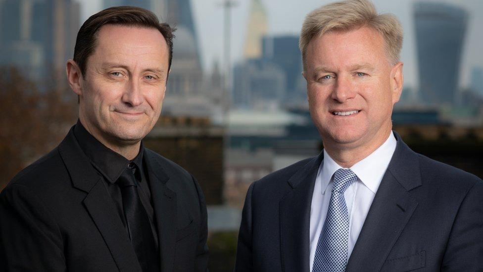 Adam is standing on the left. He has brown hair and is wearing a black suit and tie. Tom is on the right and has blonde hair and blue eyes. He is wearing a dark navy suit jacket over a white shirt and blue-spotted tie. Both are smiling to the camera.