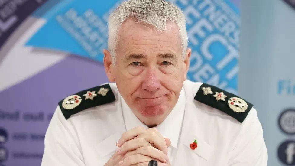 Jon Boutcher has grey hair and is leaning slightly forward in his PSNI shirt with his hands clasped in front of him