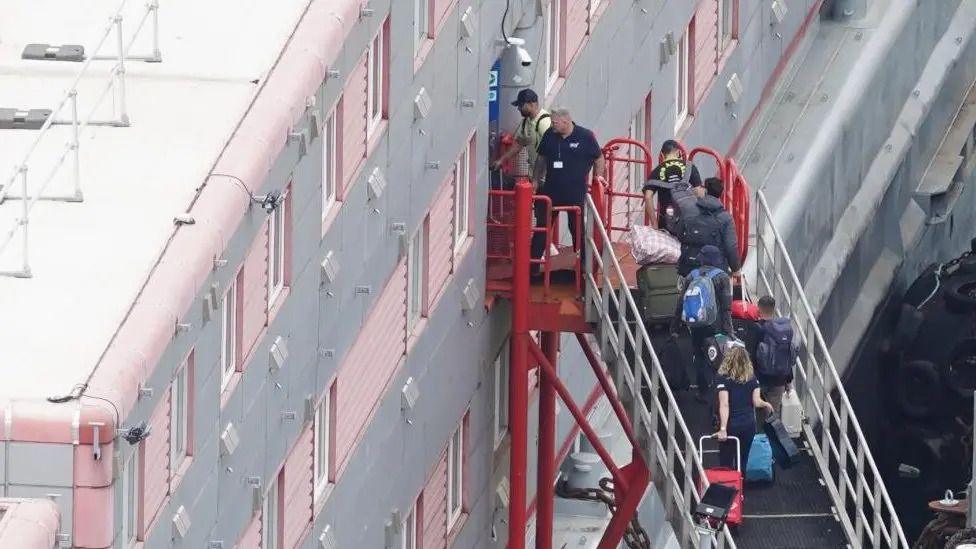 People carrying bags and suitcases walking up a ramp that leads to the entrance of a grey and red three-storey barge. 