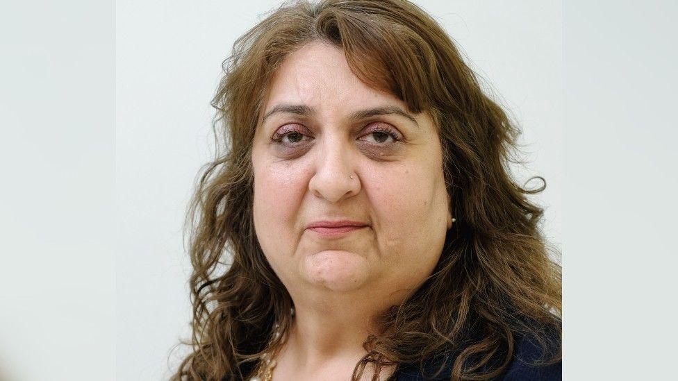 A professional headshot of a woman with curly brown hair and wearing light brown eye shadow, looking straight at the camera.