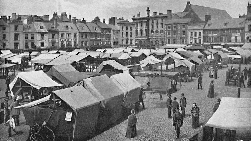 Black and white image showing canvass market stalls and women wearing long dresses and shawls as well as men in suits and hats walking between them. Tall buildings surround the market.