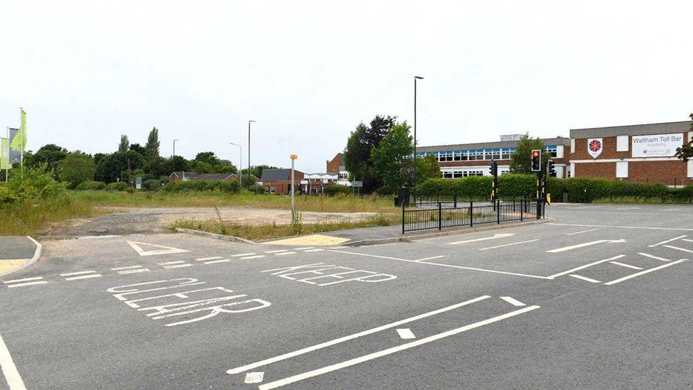 The site of the new car park overgrown with weeds located on the corner of a junction with traffic lights with the red brick, two-storey building of the school in the background