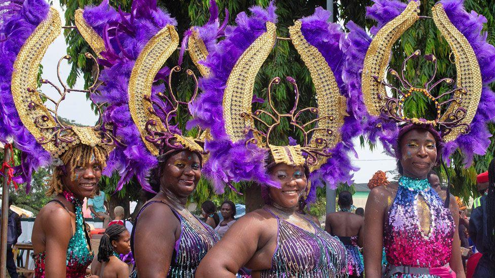 Four women in sequin outfits and purple feathered headdresses attached to manilla like horns at the Calabar carnival smile at the camera