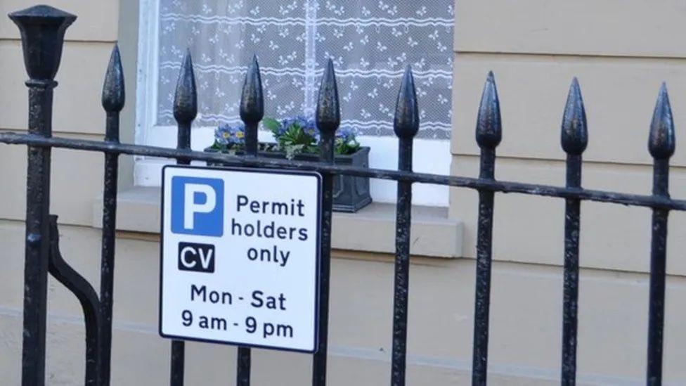 White sign displaying parking for permit holders only on a metal fence. 