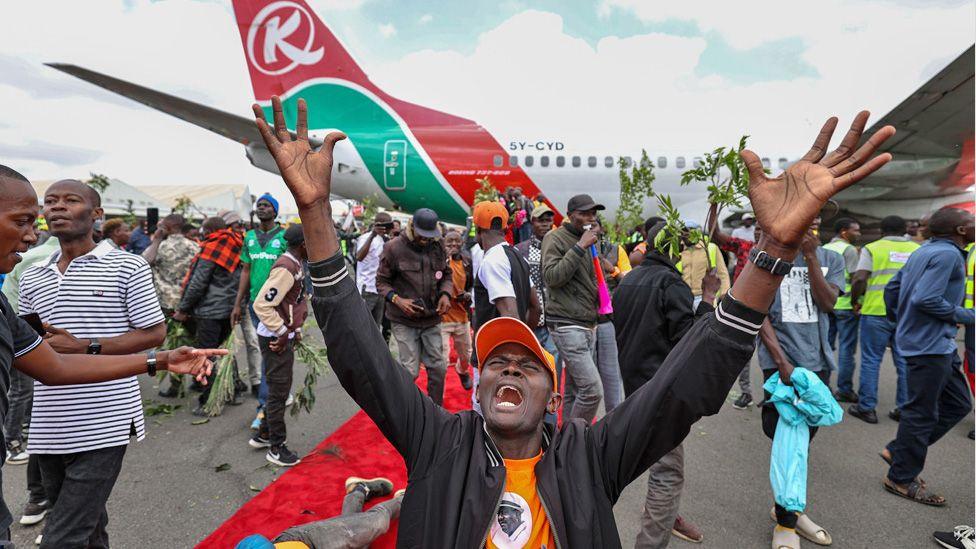 A mourner in an orange cap, orange T-shirt and black jacket with his hands in the air wailing, another fallen on a red carpet. Behind them is the plane that carried home the coffin of Raila Odinga from India - 16 October 2025