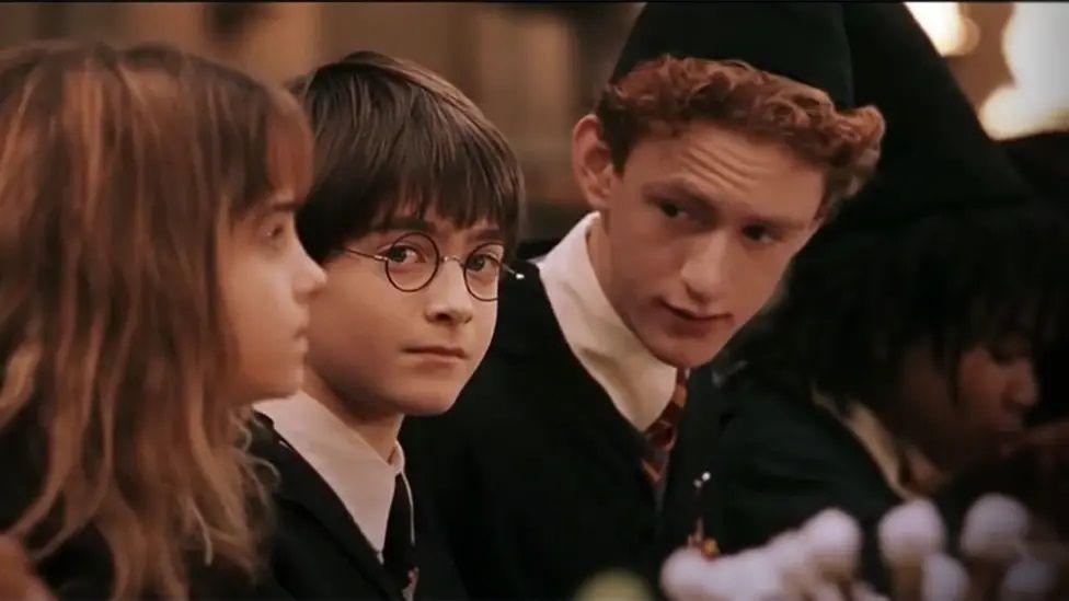 A shot from one of the early Harry Potter films, showing Hermione Granger sitting by Harry Potter and Percy Weasley. Harry looks at the camera directly from side on while Percy speaks to them both, wearing a black cap. Hermione has a fringe and brown shoulder length hair, Harry has a short bowl hair cut and wears school uniform, as does Percy who has reddish wavy short hair.