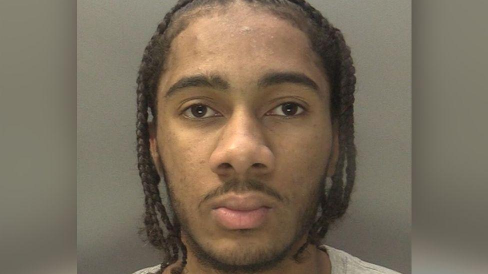 A police mugshot of a young man with chin-length dreadlocked hair.