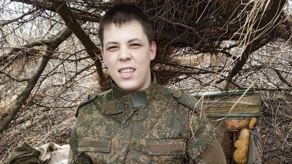 Yaroslav Lipavsky in camouflage clothing, with short, dark straight hair - he is standing in front of a man-made hole dug into the ground and some tree branches