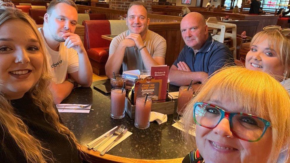 Daniel, Peter, Jean, Shayley, Mervyn and Hayley Housden sitting at a table in a restaurant looking towards the camera with cutlery and drinks on their table