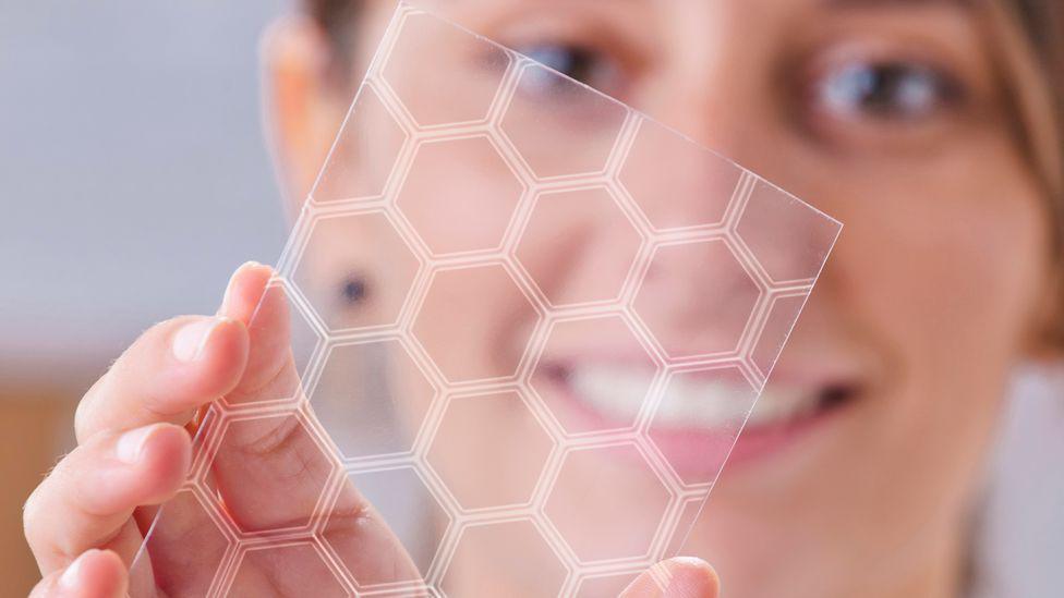 A smiling woman holds up a see-through, honeycombed shaped piece of thin material to illustrate graphene 