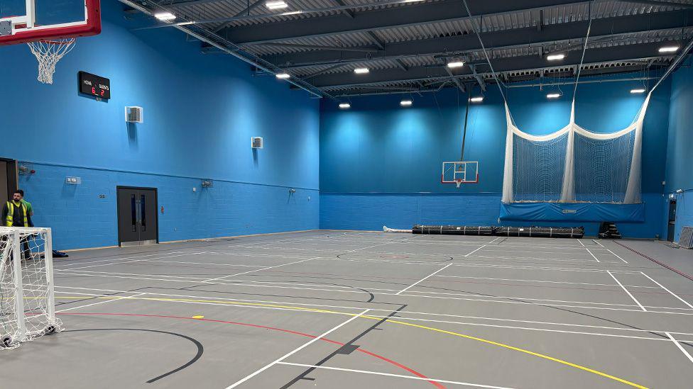 A large sports hall with blue walls and floor markings. There are five-a-side football goals and and basketball nets at either end.