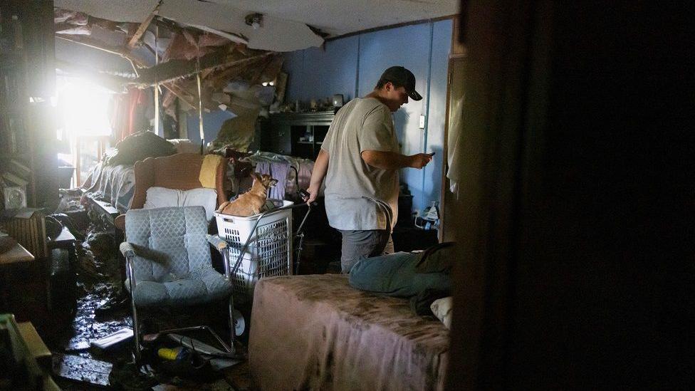 A man walks through a room which has its ceiling falling down and furniture damaged