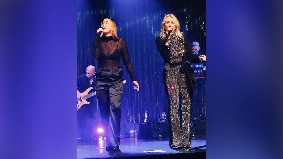 Alice Dallin singing onstage with her mum, Bananarama's Sarah Dallin. They are both wearing black, but Sarah's jumpsuit has silver sequins all over the front