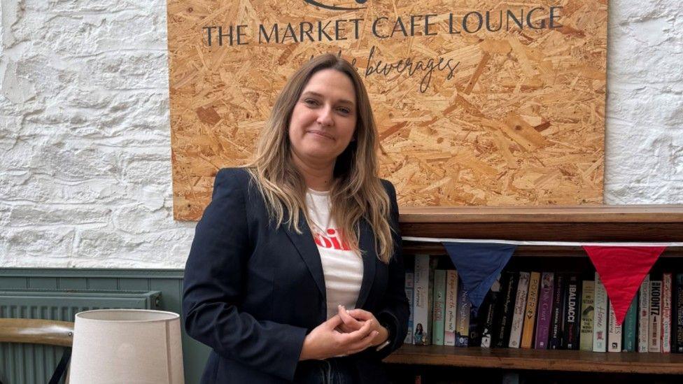 Kate leans against a book case and smiles at the camera. She is wearing a navy jacket and a white t-shirt with red writing on it. She has brown hair with blonde Ombre. Behind her is a wooden sign that reads 'The Market Café Lounge'