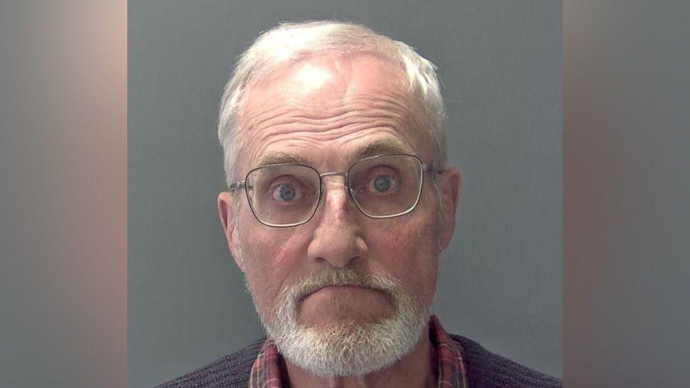 Martin Daniels is wearing glasses, and has grey/white hair and a grey/white beard and moustache. He is wearing a red tartan shirt and a ribbed brown jersey.