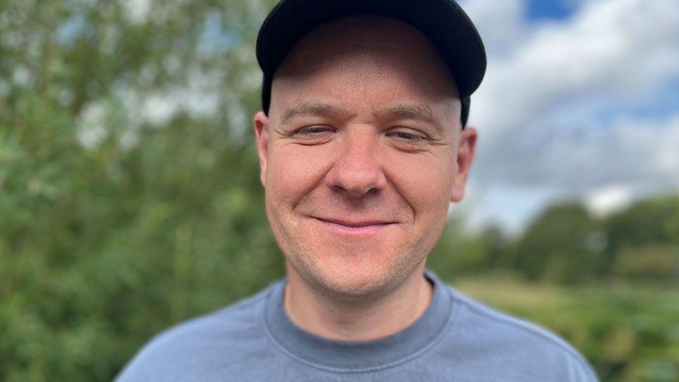 Gary Gray, who is wearing a black baseball cap and a grey T-shirt. He is starting to smile. Behind him are blurred out trees and grass.