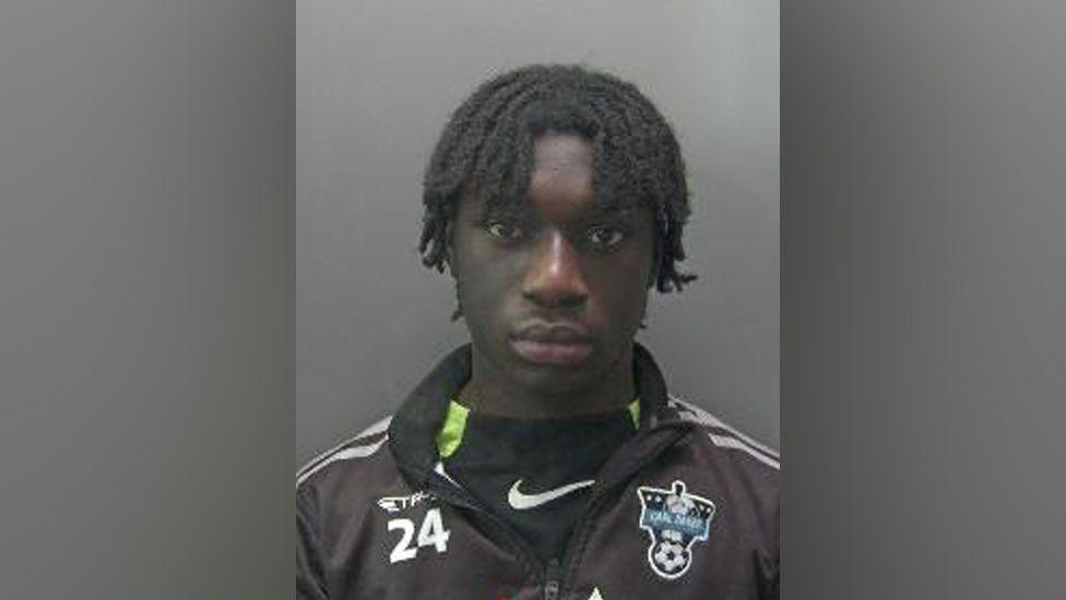 Dodou Manneh is looking at the camera in a police custody shot. He has short black hair in dreadlocks. He is wearing a black jacket and a black T-shirt.