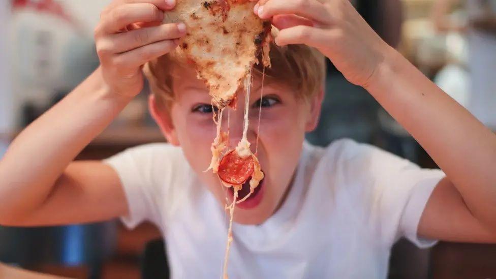 A child picks up a piece of pizza in front of their face. They look like they are making a loud noise.