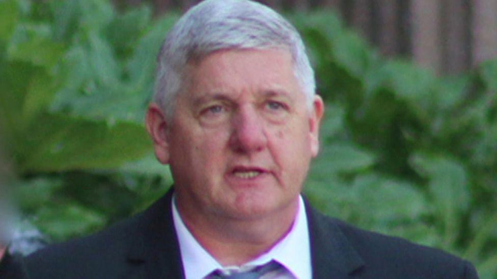 Andrew Leatherbarrow, with short grey hair and a black jacket, white shirt and blue tie, walks in front of greenery outside a court