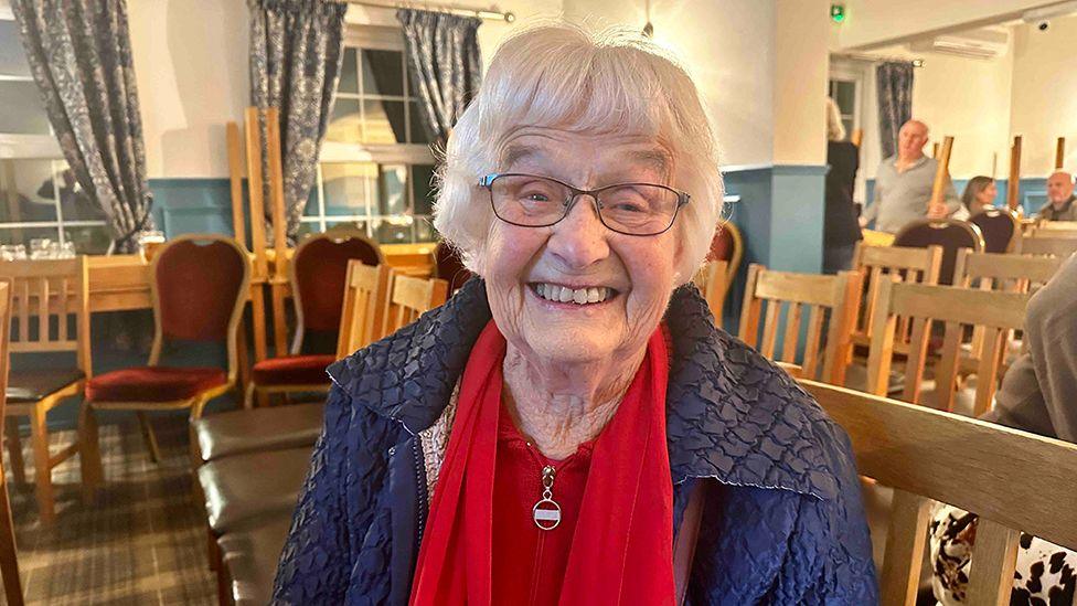 Ilene Pearson sat in the pub for the screening. She has white short hair and wears glasses and a blue coat with a red top and red scarf.