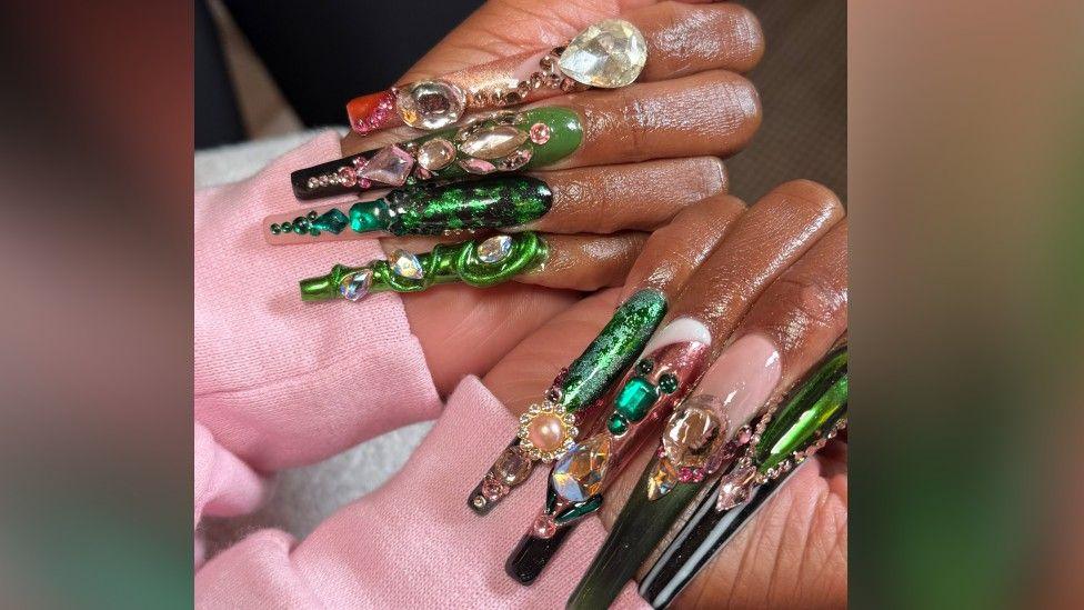 A close up of Cynthia Erivo's hands with her hands clasped shut and her nails on her palms. The sleeves of a pink hoodie can be seen. Eight nails can be seen each with green and pink designs. Some nails have a chrome metallic design on whereas others are encrusted with jewels and gems, making the design 3D. 