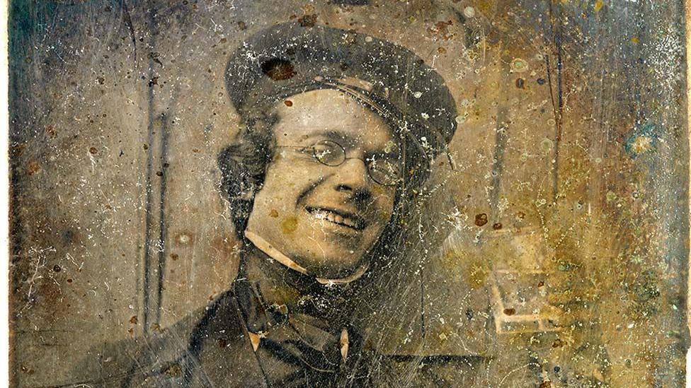 A sepia image of photography pioneer George Shaw. He is wearing Victorian clothes including a cap, shirt, jacket and neck tie. He is wearing oval glasses and smiling. 